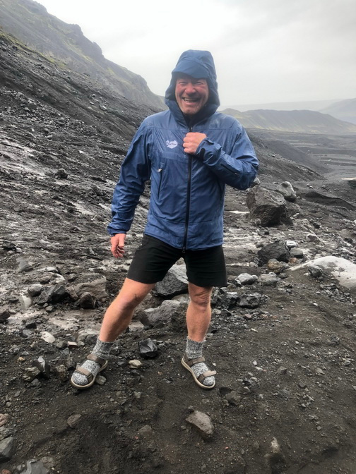Baldvin Einarsson i blå regnjakke med hette, shorts og sandaler står smilende i et steinete landskap