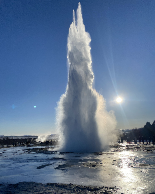 Geysir i utbrudd mot blå himmel over en snødekt slette med lav sol