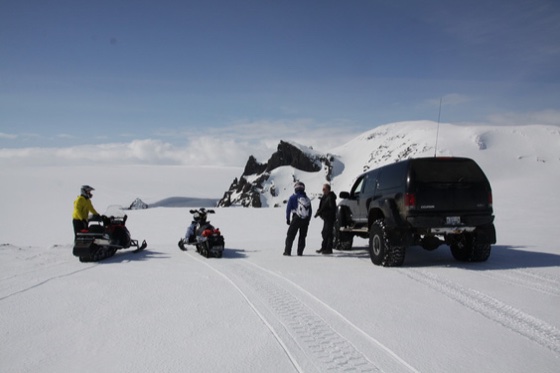 Svart Excursion parkert på snøen med to snøscootere og tre personer ved siden av