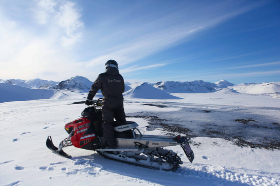 Person i kjøredress sitter på en snøscooter på et snøplatå med fjell i horisonten