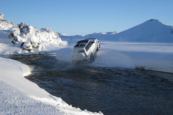 Hvit super jeep kjører opp av en elv over en snøkant med et spisst fjell bak