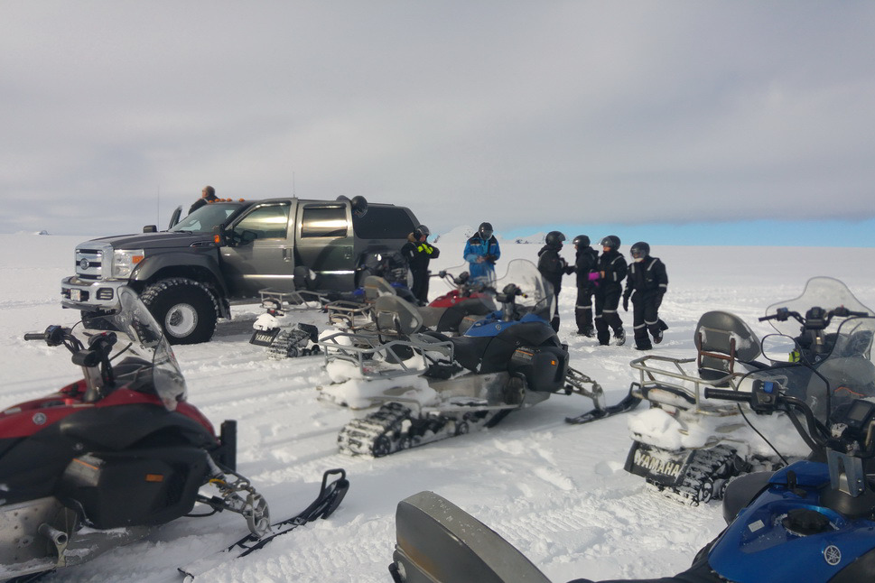 Snøscootere parkert i snøen foran en grå super jeep med en gruppe i kjøredresser