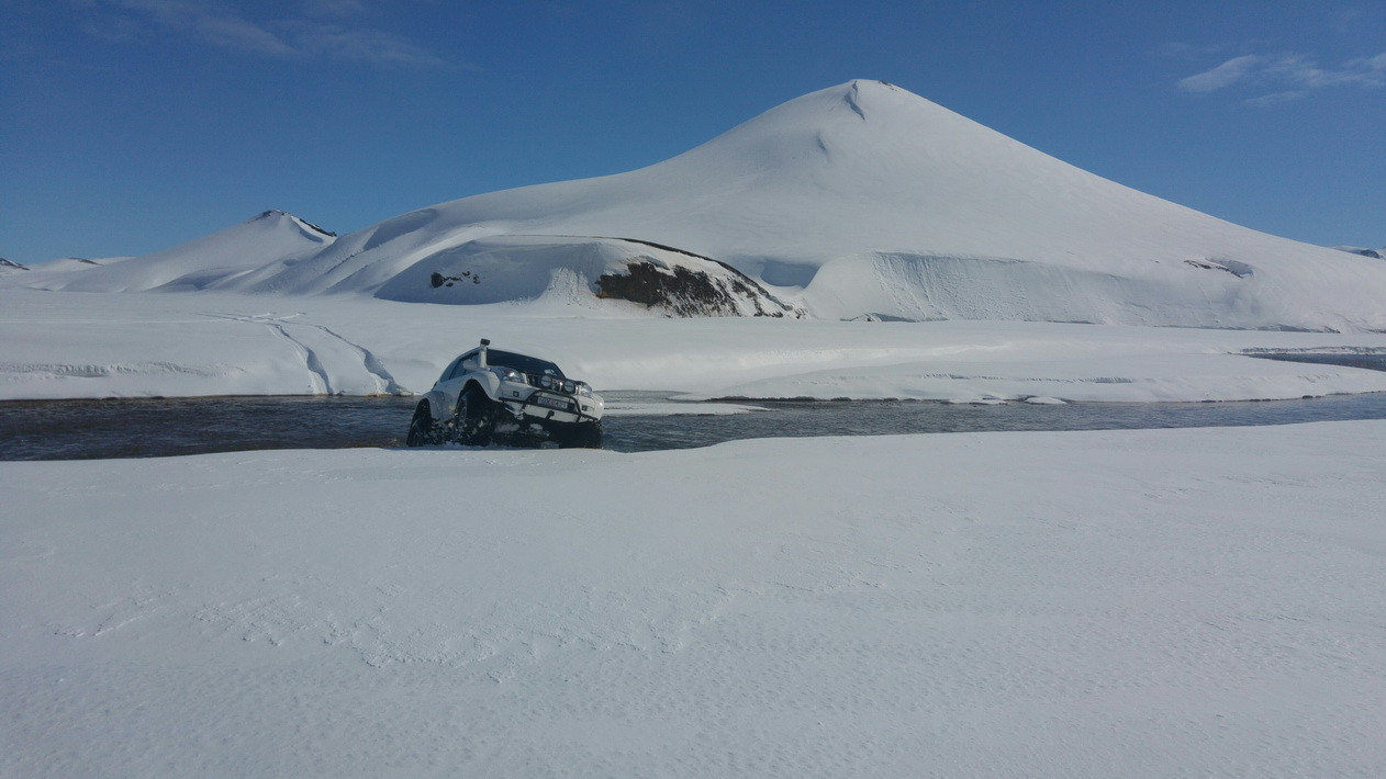 Hvit super jeep kjører gjennom en elv i et snølandskap med et snødekt fjell og blå himmel bak